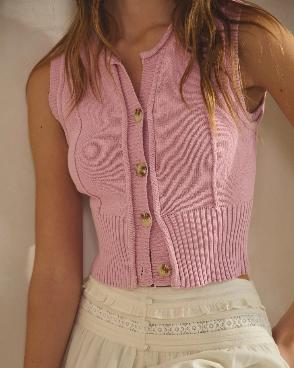 Model wearing a cropped sleeveless light pink knit vest with button front and ribbed hem styled with relaxed high-waisted bottoms.