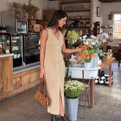 A woman wearing a beige sleeveless midi dress with a button-down front and side slit stands inside a rustic shop, holding a brown leather handbag and reaching toward flowers on a table.