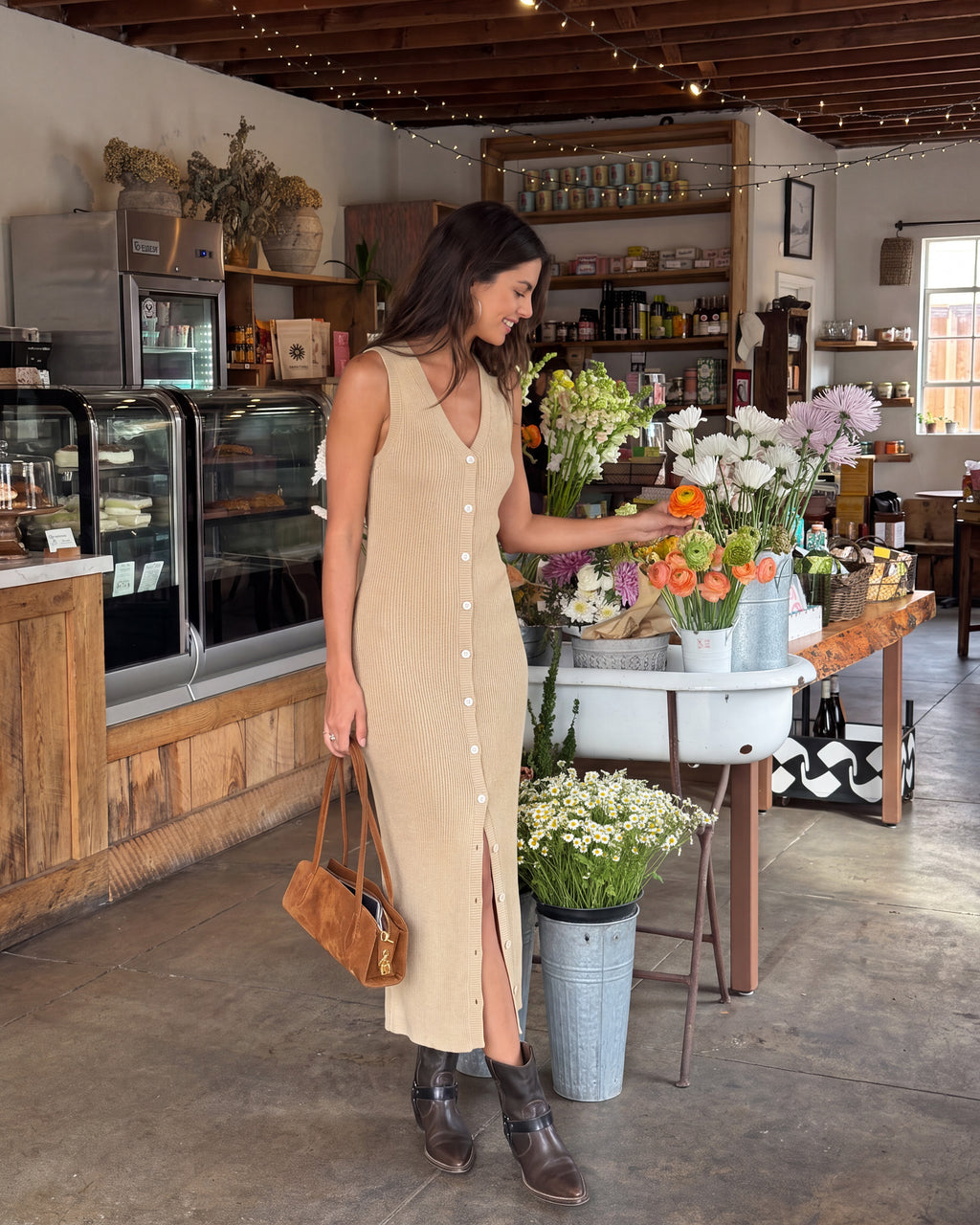 A woman wearing a beige sleeveless midi dress with a button-down front and side slit stands inside a rustic shop, holding a brown leather handbag and reaching toward flowers on a table.