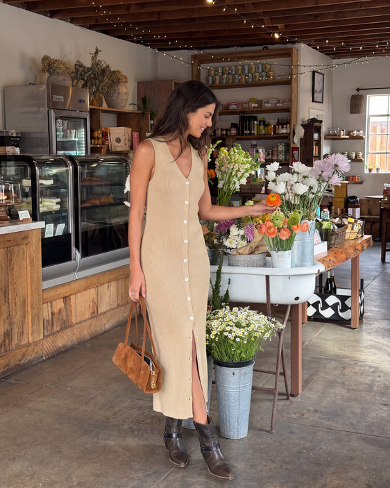 A woman wearing a beige sleeveless midi dress with a button-down front and side slit stands inside a rustic shop, holding a brown leather handbag and reaching toward flowers on a table.