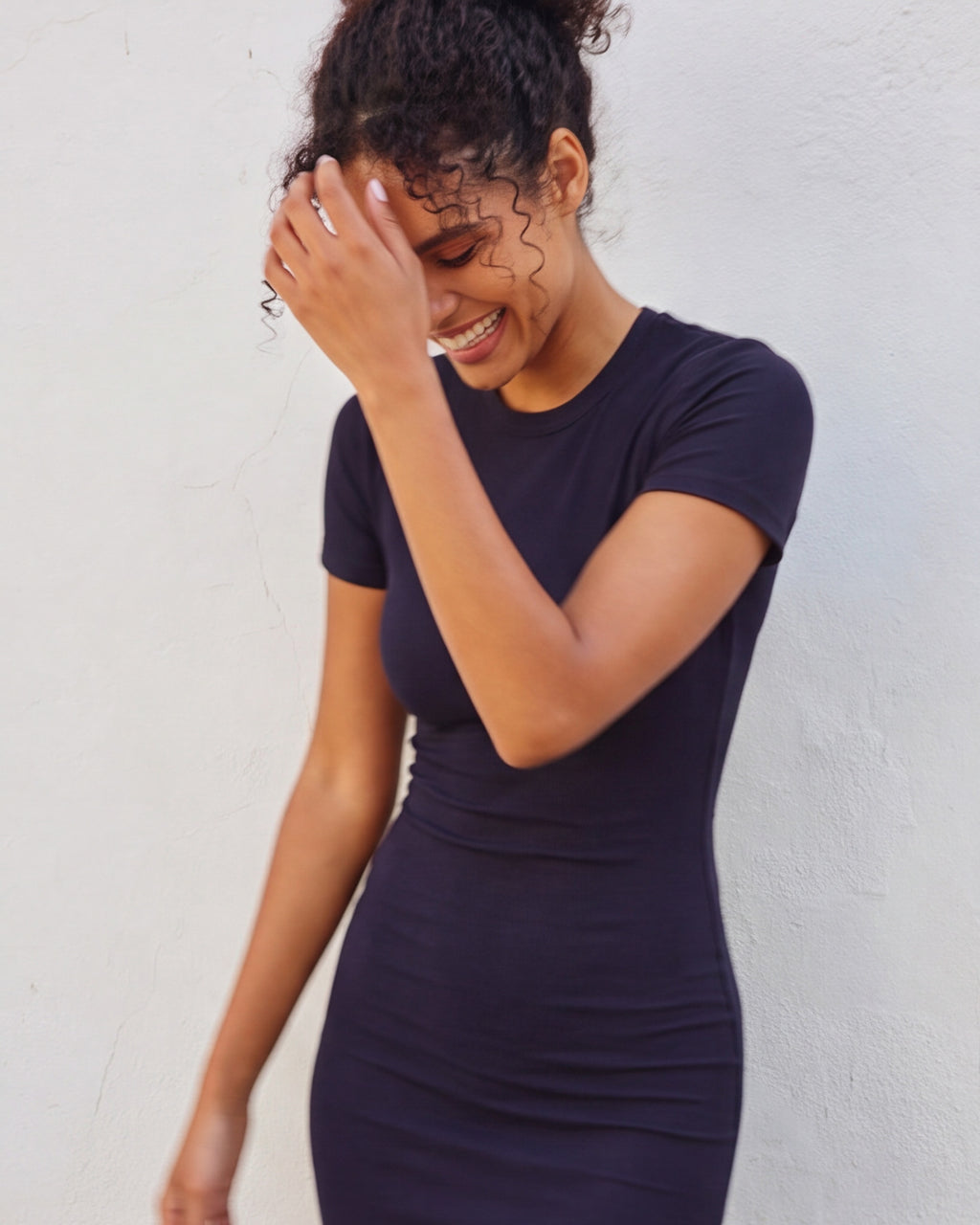 A woman wearing a dark navy blue short-sleeve bodycon dress, posing against a light-colored textured wall.