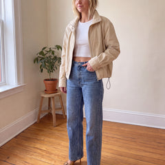 A woman stands in a room with a wood floor and cream walls, wearing a beige jacket over a white crop top, blue jeans, and beige pointed-toe heels.