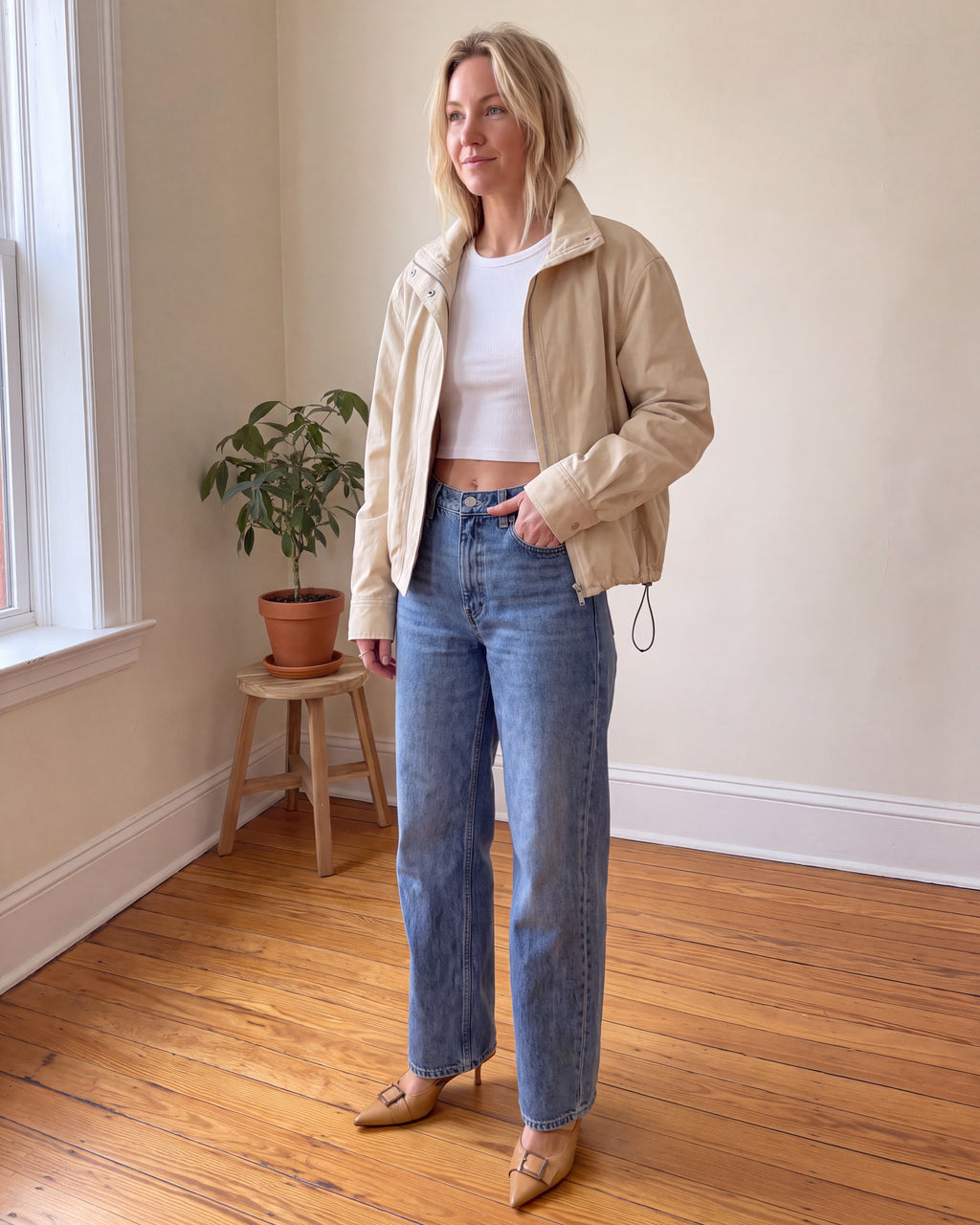 A woman stands in a room with a wood floor and cream walls, wearing a beige jacket over a white crop top, blue jeans, and beige pointed-toe heels.