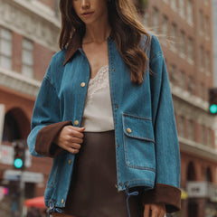 A woman wearing a blue denim jacket with brown suede-like trim on the collar and cuffs, layered over a white lace-trim top and a brown mini skirt, standing on a city street.