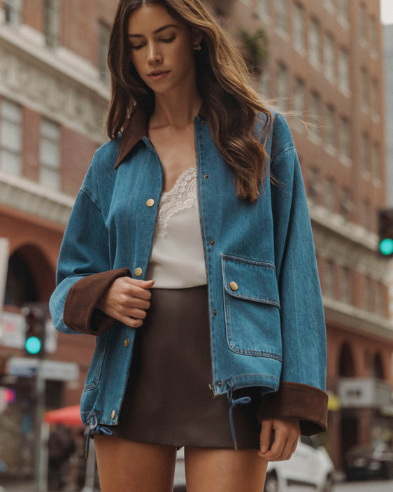 A woman wearing a blue denim jacket with brown suede-like trim on the collar and cuffs, layered over a white lace-trim top and a brown mini skirt, standing on a city street.
