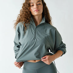A woman with curly brown hair wearing a sage green athletic half-zip pullover and matching high-waisted leggings, posing against a light gray studio background.