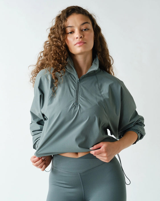 A woman with curly brown hair wearing a sage green athletic half-zip pullover and matching high-waisted leggings, posing against a light gray studio background.