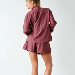 A woman shown from the back wearing a matching dusty rose athletic set consisting of a long-sleeve zip-up jacket and shorts, against a plain light gray studio background.