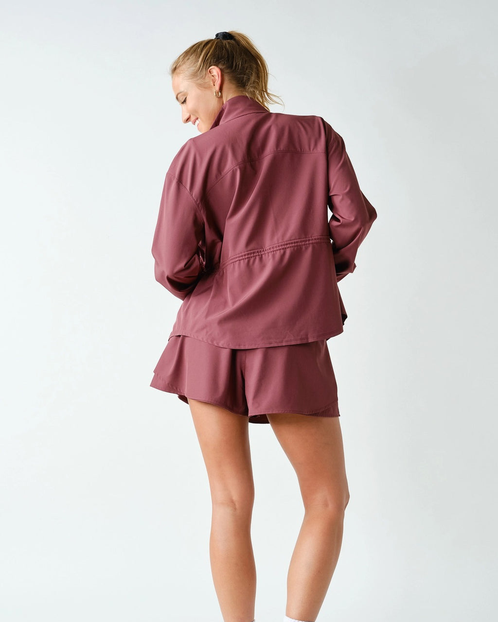 A woman shown from the back wearing a matching dusty rose athletic set consisting of a long-sleeve zip-up jacket and shorts, against a plain light gray studio background.